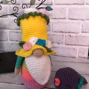 May include: A crocheted gnome with a rainbow ombre body, a yellow hat with green trim and a white beard. The gnome is standing next to a small purple crocheted bird with green wings.