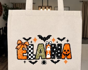 May include: A beige canvas tote bag featuring the name ELAINA in large, patterned letters. Each letter has a unique Halloween-themed design including orange spiderwebs, black and white checkers, green textures, ghosts, and a jack-o'-lantern face.
