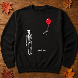 May include: A black long-sleeve sweatshirt featuring a white graphic of a skeleton looking up at a red balloon held by a detached skeletal hand. The text "Well, shit..." is printed in white below the balloon, positioned to the right of the skeleton.