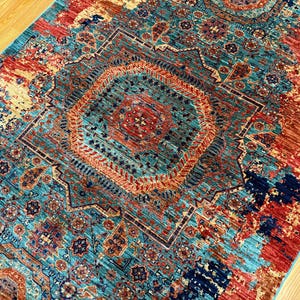 K&ouml;nnte beinhalten: Ein rechteckiger Teppich mit einem aufwendigen geometrischen Muster. Der Teppich hat ein zentrales Medaillon in T&uuml;rkis-, Rost- und Marineblaut&ouml;nen. Das Design umfasst florale und geometrische Motive, die eine Vintage- oder Boh&egrave;me-&Auml;sthetik erzeugen. Der Teppich liegt auf einem Holzboden.