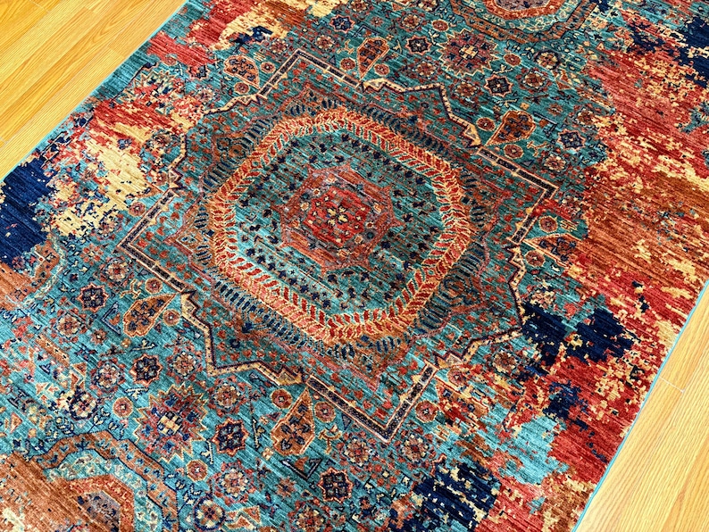 K&ouml;nnte beinhalten: Ein rechteckiger Teppich mit einem aufwendigen geometrischen Muster. Der Teppich hat ein zentrales Medaillon in T&uuml;rkis-, Rost- und Marineblaut&ouml;nen. Das Design umfasst florale und geometrische Motive, die eine Vintage- oder Boh&egrave;me-&Auml;sthetik erzeugen. Der Teppich liegt auf einem Holzboden.