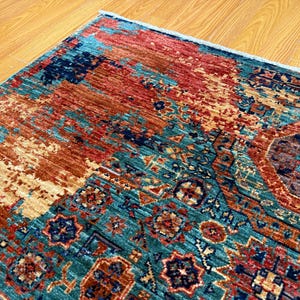 K&ouml;nnte beinhalten: Nahaufnahme eines farbenfrohen Teppichs mit einem Used-Design. Der Teppich weist eine Mischung aus T&uuml;rkis-, Rost- und Beiget&ouml;nen auf, mit komplizierten geometrischen Mustern und einem strukturierten Aussehen. Die Ecke des Teppichs ist sichtbar und zeigt die detaillierte Handwerkskunst.