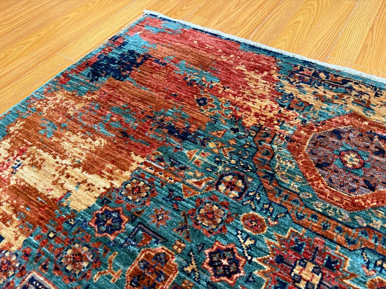 K&ouml;nnte beinhalten: Nahaufnahme eines farbenfrohen Teppichs mit einem Used-Design. Der Teppich weist eine Mischung aus T&uuml;rkis-, Rost- und Beiget&ouml;nen auf, mit komplizierten geometrischen Mustern und einem strukturierten Aussehen. Die Ecke des Teppichs ist sichtbar und zeigt die detaillierte Handwerkskunst.