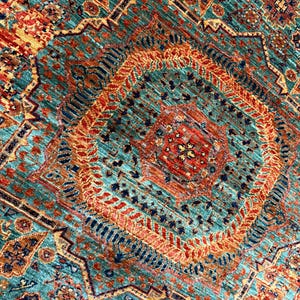 K&ouml;nnte beinhalten: Nahaufnahme eines gemusterten Teppichs mit einem zentralen Rautenmuster. Der Teppich hat einen t&uuml;rkisfarbenen Hintergrund mit komplizierten geometrischen Mustern in Orange, Rot und Marineblau. Die Textur des Teppichs ist sichtbar.