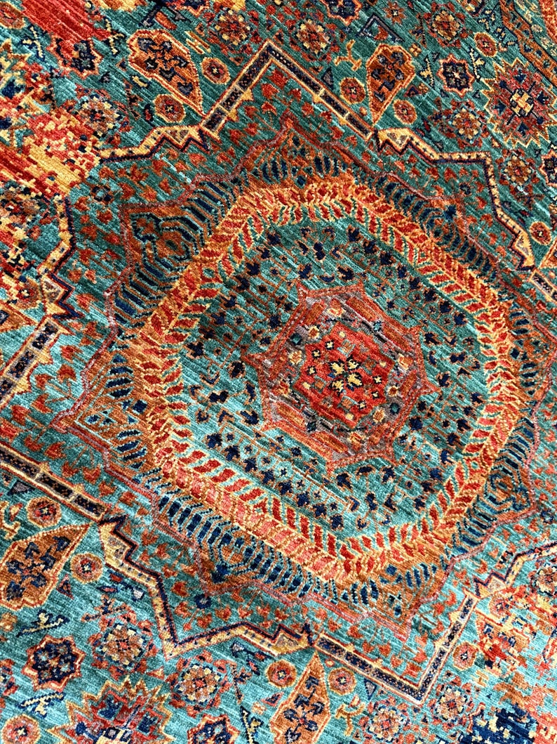 K&ouml;nnte beinhalten: Nahaufnahme eines gemusterten Teppichs mit einem zentralen Rautenmuster. Der Teppich hat einen t&uuml;rkisfarbenen Hintergrund mit komplizierten geometrischen Mustern in Orange, Rot und Marineblau. Die Textur des Teppichs ist sichtbar.