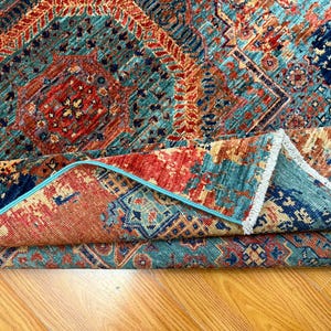 K&ouml;nnte beinhalten: Gefalteter Teppich mit einem komplexen geometrischen Muster. Der Teppich weist eine Kombination aus T&uuml;rkis, Rot und Marineblau auf. Die R&auml;nder sind mit einem hellblauen Rand versehen. Der Teppich liegt auf einer Holzoberfl&auml;che.