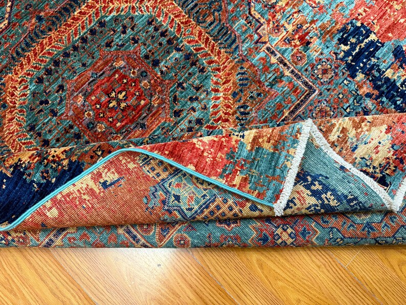 K&ouml;nnte beinhalten: Gefalteter Teppich mit einem komplexen geometrischen Muster. Der Teppich weist eine Kombination aus T&uuml;rkis, Rot und Marineblau auf. Die R&auml;nder sind mit einem hellblauen Rand versehen. Der Teppich liegt auf einer Holzoberfl&auml;che.