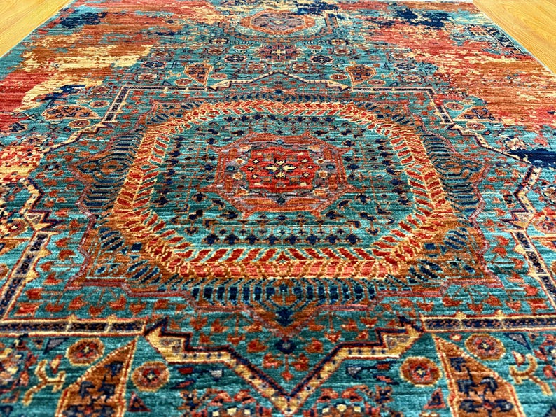 K&ouml;nnte beinhalten: Ein gro&szlig;er, rechteckiger Teppich mit einem aufwendigen, verwitterten Muster. Der Teppich hat ein zentrales Medaillon-Design in T&uuml;rkis, Orange und Marineblau. Die Gesamt&auml;sthetik ist eine Mischung aus traditionellen und modernen Stilen.