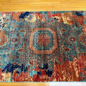 K&ouml;nnte beinhalten: Ein rechteckiger Teppich mit einem Used-Look-Design im Vintage-Stil. Der Teppich hat ein zentrales Medaillon in T&uuml;rkis-, Orange- und Rott&ouml;nen, umgeben von einem Rand in &auml;hnlichen Farben. Das Muster des Teppichs hat ein verblasstes Aussehen.