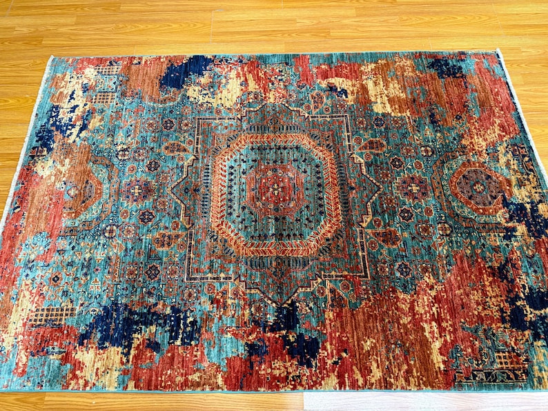 K&ouml;nnte beinhalten: Ein rechteckiger Teppich mit einem Used-Look-Design im Vintage-Stil. Der Teppich hat ein zentrales Medaillon in T&uuml;rkis-, Orange- und Rott&ouml;nen, umgeben von einem Rand in &auml;hnlichen Farben. Das Muster des Teppichs hat ein verblasstes Aussehen.