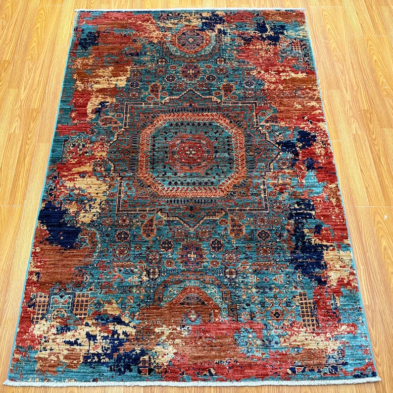 K&ouml;nnte beinhalten: Ein rechteckiger Teppich mit einem aufwendigen, verwitterten Design. Der Teppich zeigt ein zentrales Medaillon und einen Rand in T&uuml;rkis, Rot, Rost und Marineblau. Das Muster des Teppichs hat ein Vintage-Aussehen.