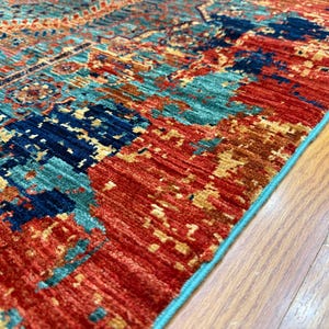 K&ouml;nnte beinhalten: Nahaufnahme eines farbenfrohen Teppichs mit einem Used-Look-Design. Der Teppich weist eine Mischung aus roten, teal- und marineblauen Farbt&ouml;nen mit einem hellblauen Rand auf. Der Teppich liegt auf einem Holzboden.