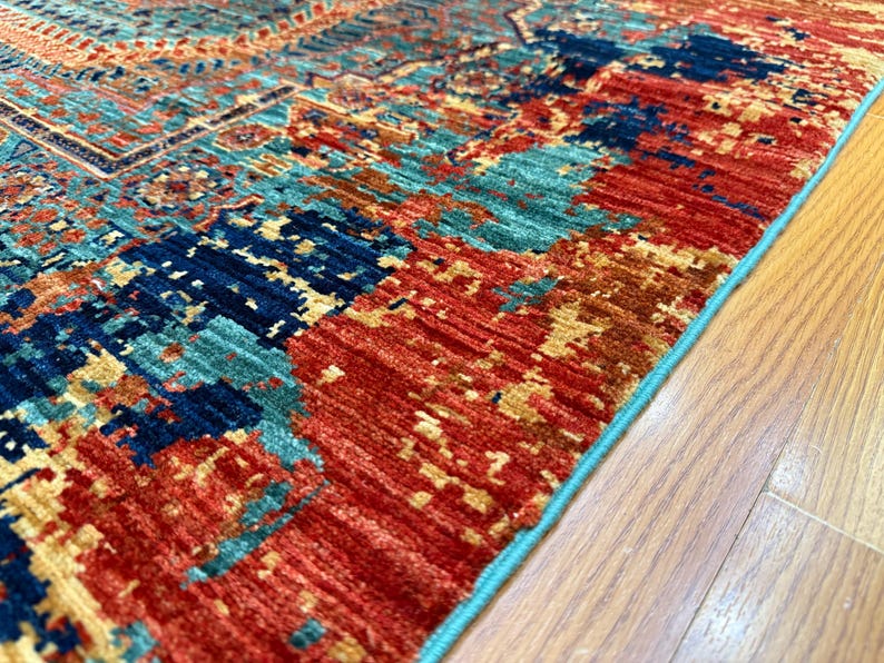 K&ouml;nnte beinhalten: Nahaufnahme eines farbenfrohen Teppichs mit einem Used-Look-Design. Der Teppich weist eine Mischung aus roten, teal- und marineblauen Farbt&ouml;nen mit einem hellblauen Rand auf. Der Teppich liegt auf einem Holzboden.