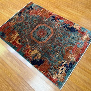 K&ouml;nnte beinhalten: Rechteckiger Teppich mit einem verwitterten, Vintage-Look. Der Teppich hat ein zentrales Medaillon-Design in T&uuml;rkis-, Rot- und Braunt&ouml;nen, mit einem Rand in &auml;hnlichen Farben. Er liegt auf einem hellen Holzboden.