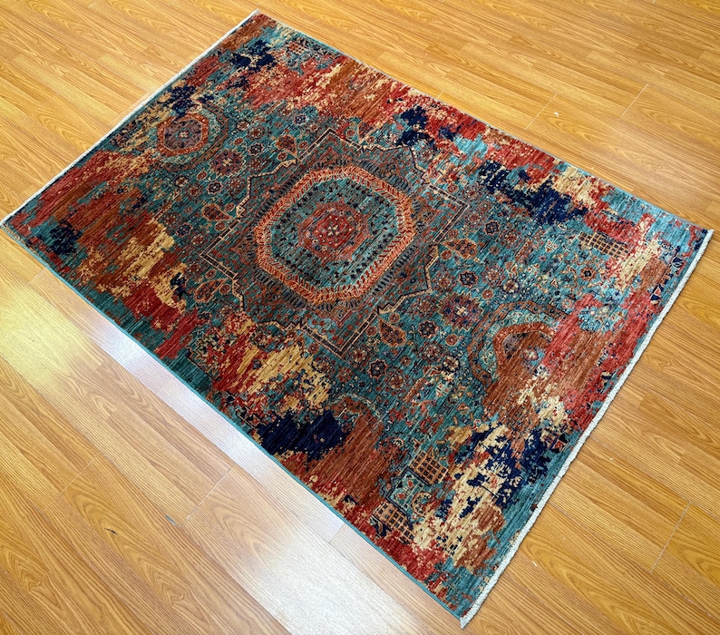 K&ouml;nnte beinhalten: Rechteckiger Teppich mit einem verwitterten, Vintage-Look. Der Teppich hat ein zentrales Medaillon-Design in T&uuml;rkis-, Rot- und Braunt&ouml;nen, mit einem Rand in &auml;hnlichen Farben. Er liegt auf einem hellen Holzboden.