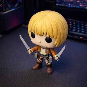 May include: A Funko Pop figure of a character with blonde hair, wearing a brown jacket, white shirt, and brown boots, holding two silver knives. The figure has a detailed design and is posed on a dark surface.