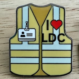 May include: A yellow safety vest with a black and white striped pattern. The vest has a black name tag with a white silhouette of a person and three white lines. The vest also has a black I heart LDC graphic with a red heart.