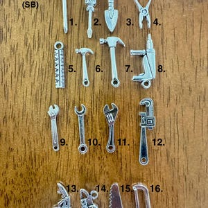 May include: A set of 16 silver and black tool charms, including a screwdriver, shovel, pliers, hammer, ruler, drill, wrench, and saw. The charms are numbered 1 through 16.