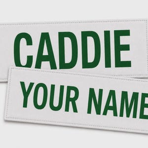 May include: Two rectangular white fabric patches with stitched borders. The top patch features the word CADDIE in bold dark green capital letters. The bottom patch features the words YOUR NAME in bold dark green capital letters.