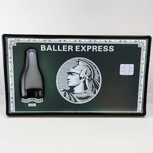 Op de afbeelding: Een zwart en groen metalen bord met de tekst "BALLER EXPRESS" in wit. Het bord heeft een gestileerde afbeelding van een persoon met een helm en een chipkaart.