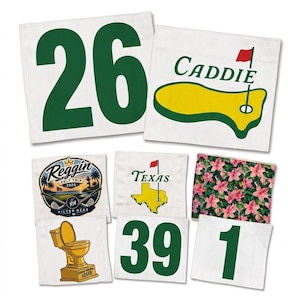 May include: A collection of white fabric golf flags featuring various designs including the number 26 in green, a yellow golf green with the word Caddie, a Reggin Golf Trip 2026 logo, a Texas map with a flag, pink flowers, a gold toilet trophy, 39, and 1.