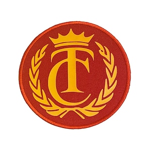 May include: A round, red patch with a gold crown, the letters "TF", and a laurel wreath design. The patch is likely for clothing or accessories, and the design is centered on a white background.