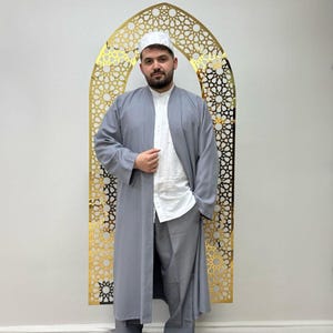 Puede incluir: Un hombre con una túnica gris claro abierta sobre una camisa blanca y pantalones grises a juego. También lleva una gorra blanca. El fondo presenta un arco decorativo dorado con un intrincado patrón geométrico.