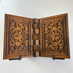 Puede incluir: Un atril de madera con intrincados dise&ntilde;os tallados. El atril est&aacute; hecho de madera marr&oacute;n oscuro y presenta un dise&ntilde;o con bisagras con una serie de bloques de madera que conectan los dos lados. Los detalles tallados son visibles en la parte frontal de cada panel.
