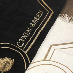 May include: Black and cream-colored prayer rugs with gold embroidered details. The black rug features the text "CANIM BABAM" in gold script and a gold tulip design. The cream rug has a geometric gold pattern and fringe.