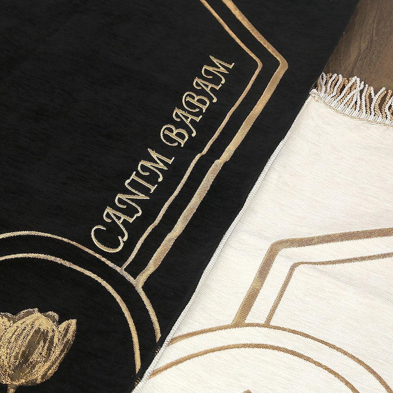 May include: Black and cream-colored prayer rugs with gold embroidered details. The black rug features the text "CANIM BABAM" in gold script and a gold tulip design. The cream rug has a geometric gold pattern and fringe.