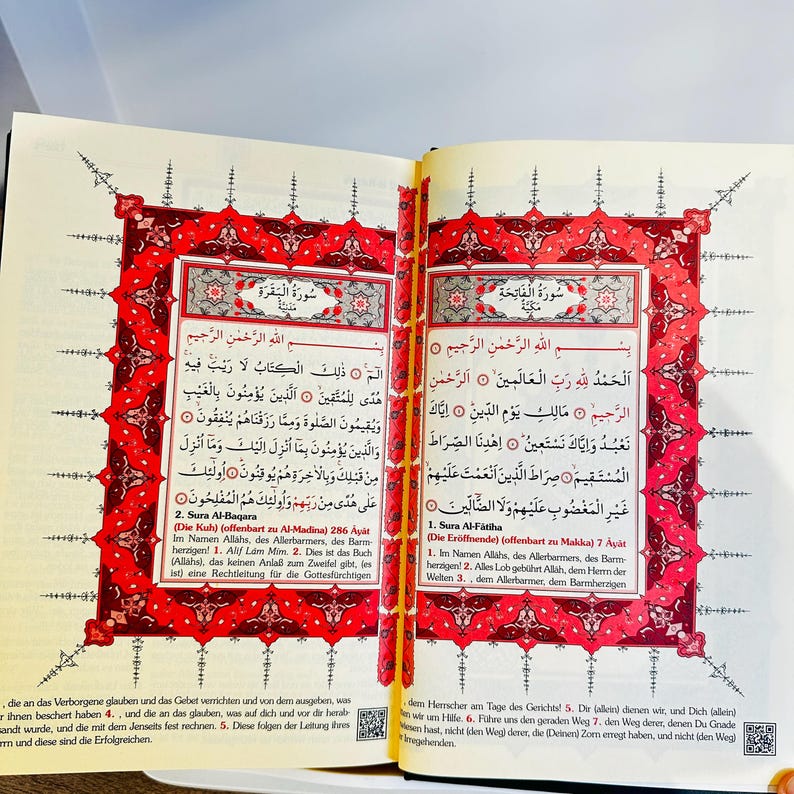 May include: Open book with Arabic text and ornate red and gold borders. The pages display verses from the Quran, with Arabic script and translations. The book is open to two facing pages.