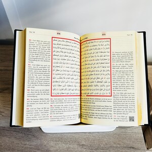 May include: An open book with Arabic text, likely a religious text, resting on a white surface. The pages are yellowed, and the text is in black. The book is bound in black.