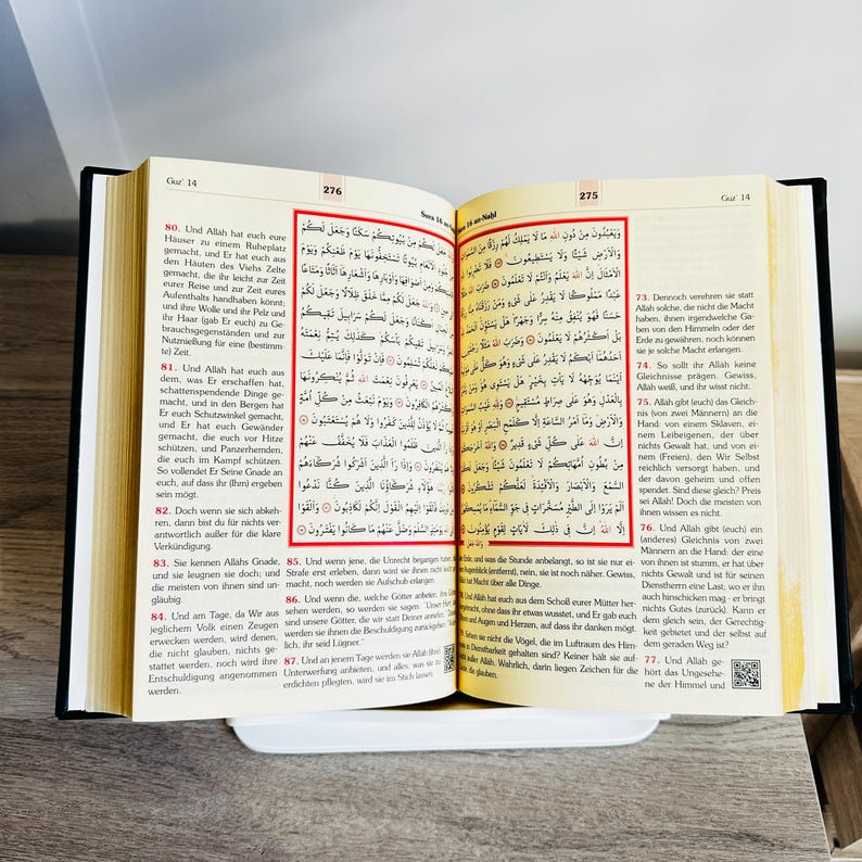 May include: An open book with Arabic text, likely a religious text, resting on a white surface. The pages are yellowed, and the text is in black. The book is bound in black.