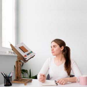 K&ouml;nnte beinhalten: Ein B&uuml;cherst&auml;nder aus Holz mit aufwendigen Schnitzereien, der ein Buch offen auf einer Seite mit einer Farbkarte h&auml;lt. Eine Person sitzt an einem Schreibtisch mit einem Notizbuch und einer rosa Tasse.