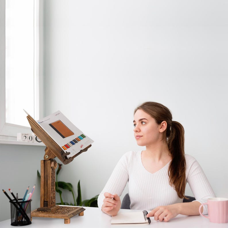 K&ouml;nnte beinhalten: Ein B&uuml;cherst&auml;nder aus Holz mit aufwendigen Schnitzereien, der ein Buch offen auf einer Seite mit einer Farbkarte h&auml;lt. Eine Person sitzt an einem Schreibtisch mit einem Notizbuch und einer rosa Tasse.