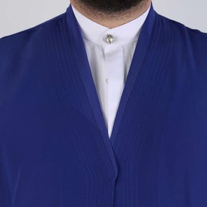 Op de afbeelding: Een close-up van een blauw kledingstuk met een V-hals, gedragen over een wit overhemd met kraag. De blauwe stof heeft subtiele verticale stiksels. Een zilveren knoop siert de kraag van het overhemd. De stijl suggereert formele of semi-formele kleding.