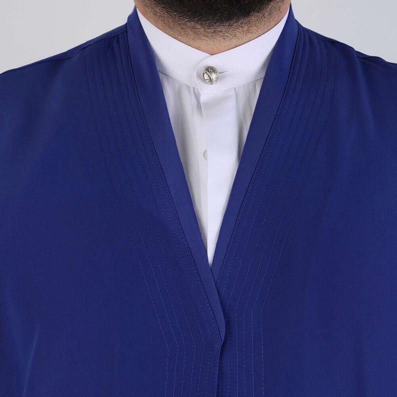 Op de afbeelding: Een close-up van een blauw kledingstuk met een V-hals, gedragen over een wit overhemd met kraag. De blauwe stof heeft subtiele verticale stiksels. Een zilveren knoop siert de kraag van het overhemd. De stijl suggereert formele of semi-formele kleding.