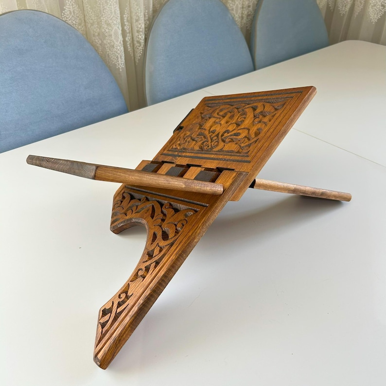 Puede incluir: Un atril de madera con intrincadas tallas florales. El atril est&aacute; hecho de madera de color marr&oacute;n claro y presenta una superficie inclinada para sostener un libro. Tiene una barra de soporte y una base con recortes decorativos.