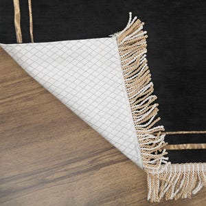 May include: Black rug with gold stripes and fringe. The rug's underside is white with a diamond pattern. The rug is partially folded back, revealing the underside and the wooden floor.