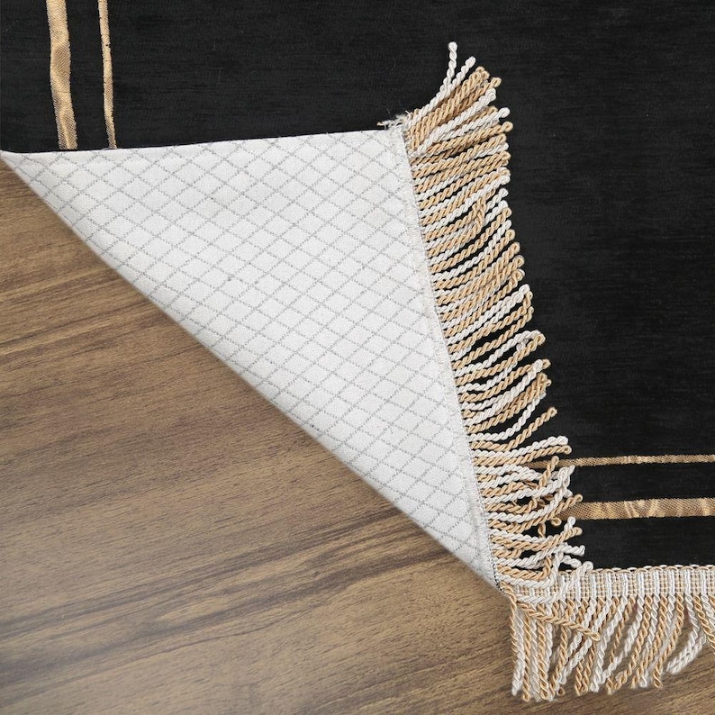 May include: Black rug with gold stripes and fringe. The rug's underside is white with a diamond pattern. The rug is partially folded back, revealing the underside and the wooden floor.