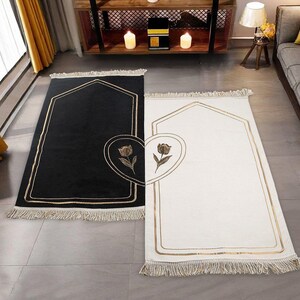 May include: Two prayer rugs, one black and one white, with gold trim and a floral heart design. Each rug has a rectangular shape with an arched top and fringe along the bottom edge. The rugs are placed on a gray floor.