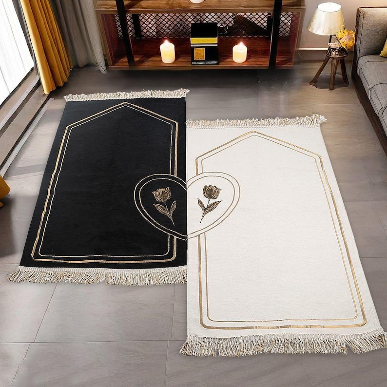 May include: Two prayer rugs, one black and one white, with gold trim and a floral heart design. Each rug has a rectangular shape with an arched top and fringe along the bottom edge. The rugs are placed on a gray floor.