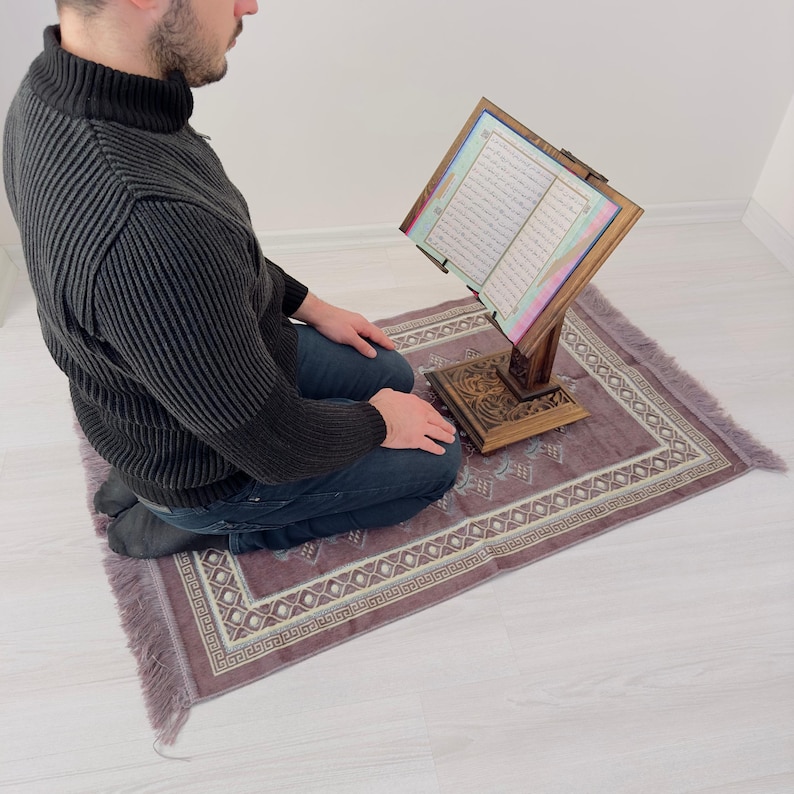 K&ouml;nnte beinhalten: Eine Person kniet auf einem Gebetsteppich mit einem h&ouml;lzernen Buchst&auml;nder, der ein offenes Buch mit arabischem Text h&auml;lt. Der Teppich ist lila mit einem wei&szlig;en und goldenen geometrischen Muster.