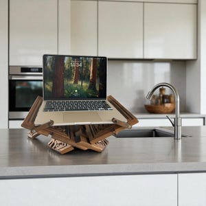Puede incluir: Un soporte de madera para port&aacute;til con un dise&ntilde;o decorativo tallado. El soporte sostiene un port&aacute;til con una escena de bosque en la pantalla. El soporte es de madera marr&oacute;n claro y est&aacute; sobre una encimera gris.