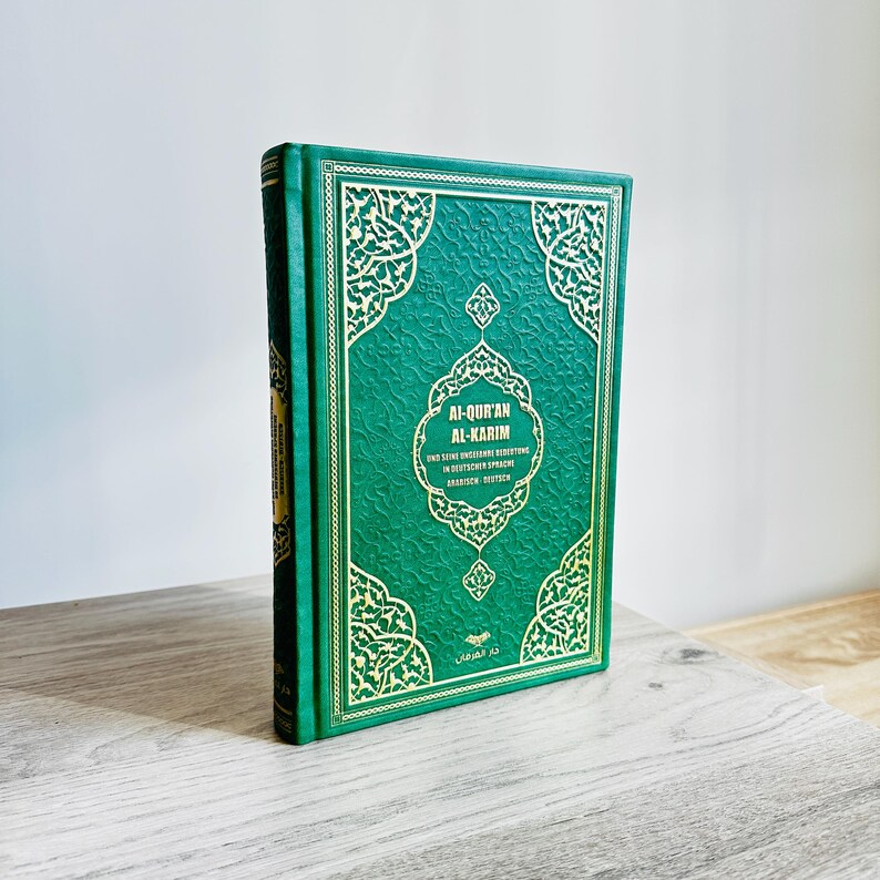 May include: A hardcover book titled "Al-Qur'an Al-Karim" in green with gold decorative accents. The book is standing upright on a wooden surface. The cover features ornate patterns and text in both Arabic and German.