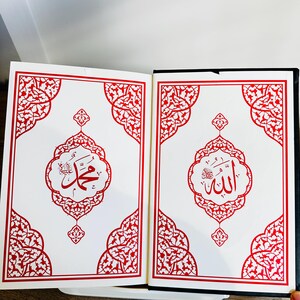 May include: Open book with white pages featuring red ornate designs and Arabic calligraphy. The left page displays the name "Muhammad" in red script within a decorative frame. The right page shows the name "Allah" in red script.