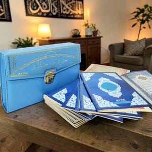 Può includere: Una valigetta azzurra in similpelle con dettagli dorati e chiusura dorata, accanto a diversi libri aperti con copertine blu e scritte dorate. I libri sono disposti su un tavolo di legno, con testo visibile sulle pagine. La valigetta riporta la scritta "AYFA".