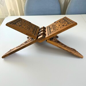 Puede incluir: Un atril de madera con intrincadas tallas florales. El atril est&aacute; hecho de madera de tono medio y tiene un dise&ntilde;o plegable. El atril est&aacute; abierto, mostrando las tallas detalladas en la parte superior e inferior. Est&aacute; dise&ntilde;ado para sostener un libro.