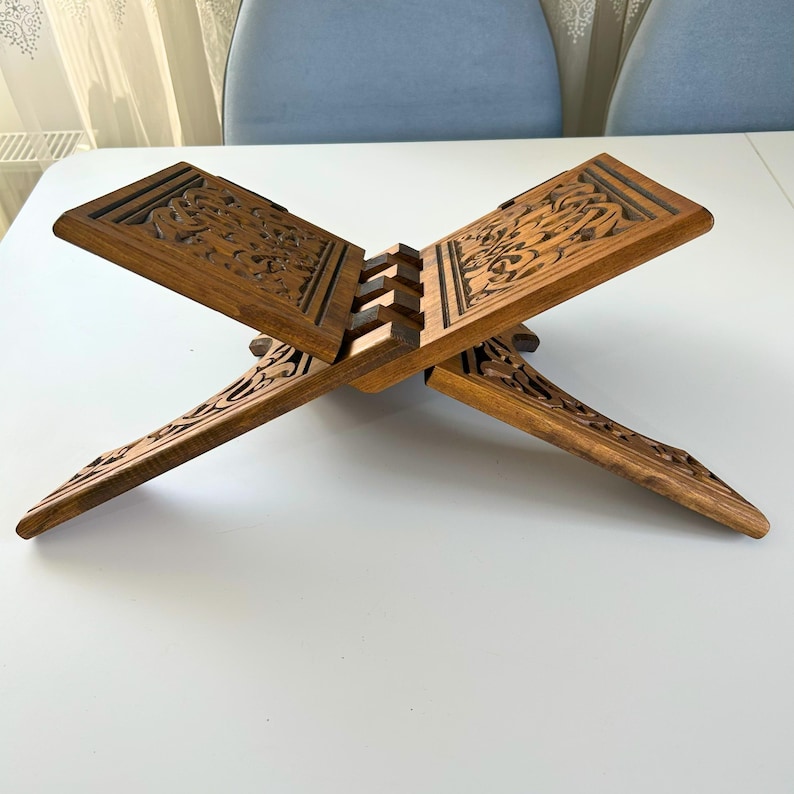 Puede incluir: Un atril de madera con intrincadas tallas florales. El atril est&aacute; hecho de madera de tono medio y tiene un dise&ntilde;o plegable. El atril est&aacute; abierto, mostrando las tallas detalladas en la parte superior e inferior. Est&aacute; dise&ntilde;ado para sostener un libro.