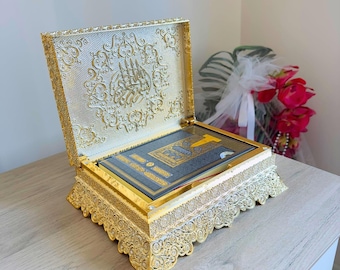 May include: A gold-colored decorative box with ornate detailing, open to reveal a book with gold accents. The box features intricate patterns and a hinged lid. The box is sitting on a light-colored wooden surface.